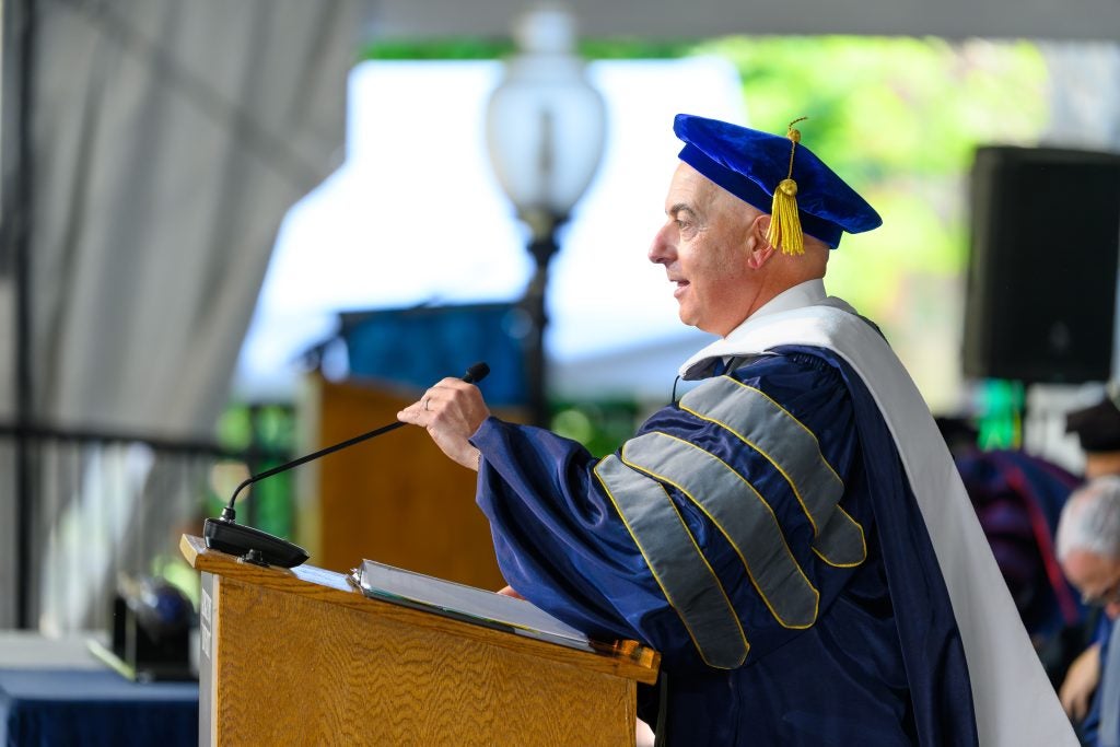 A man in a graduation gown with a blue graduation gown speaks behind a podium