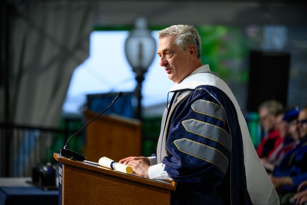 A gray-haired man in a graduation gown stands behind a podium