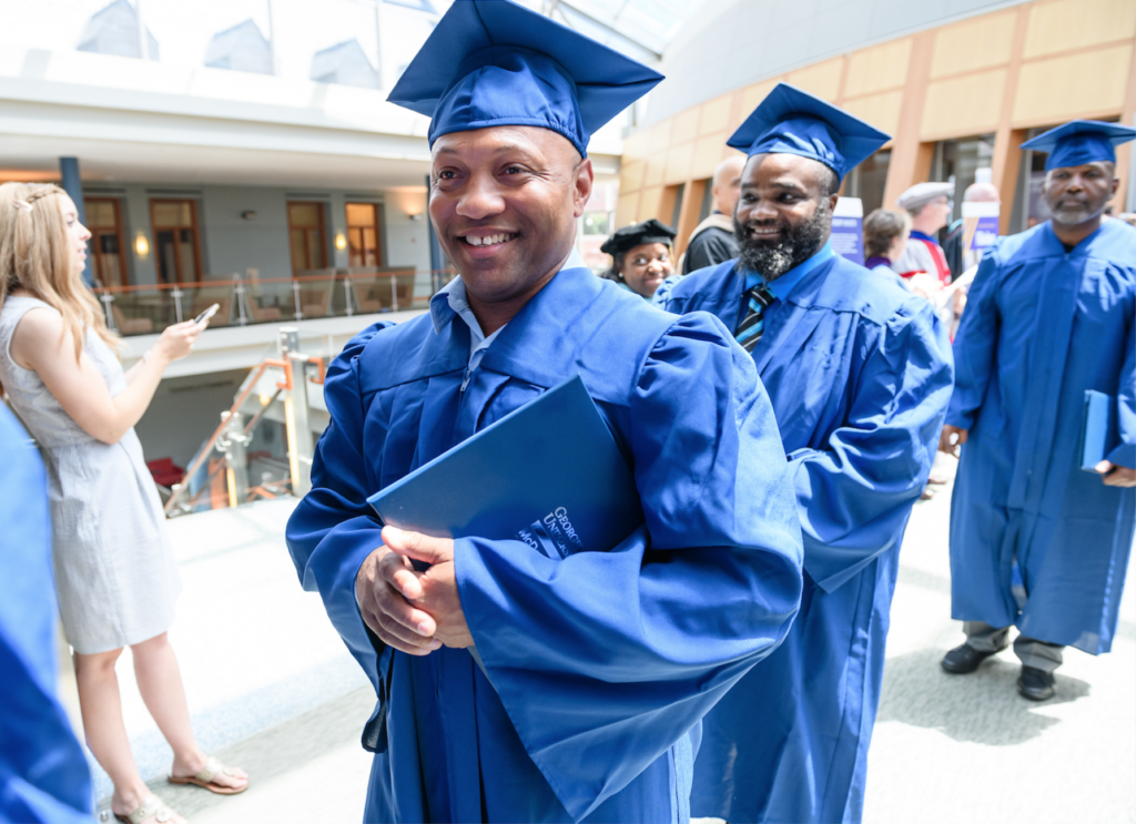 A man in a blue graduation cap and gown smiles as he holds a certificate