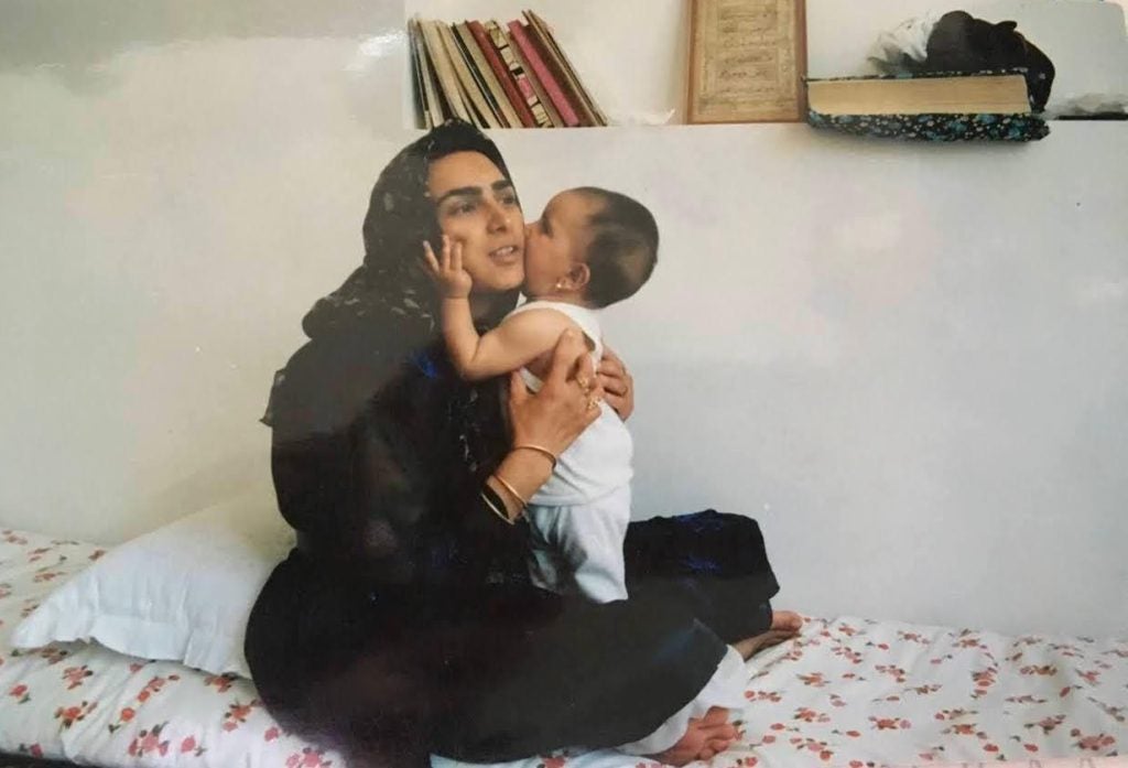 A mother in a hijab holds her baby on a bed. Behind her are books