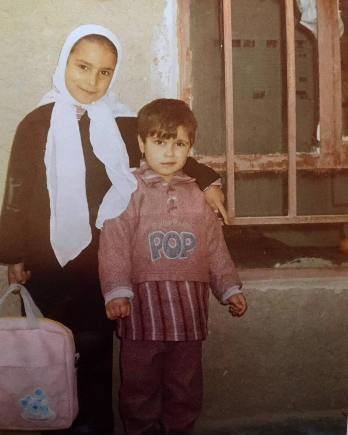 A photo of two children in Afghanistan. One wears a white hijab and carries a backpack; the other has a pink shirt that says "POP"