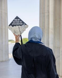 A woman with a hijab holds up a graduation cap that says on the top "For My Sisters in Afghanistan"