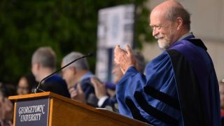 Dean William Treanor applauds behind a Georgetown University podium wearing academic regalia.