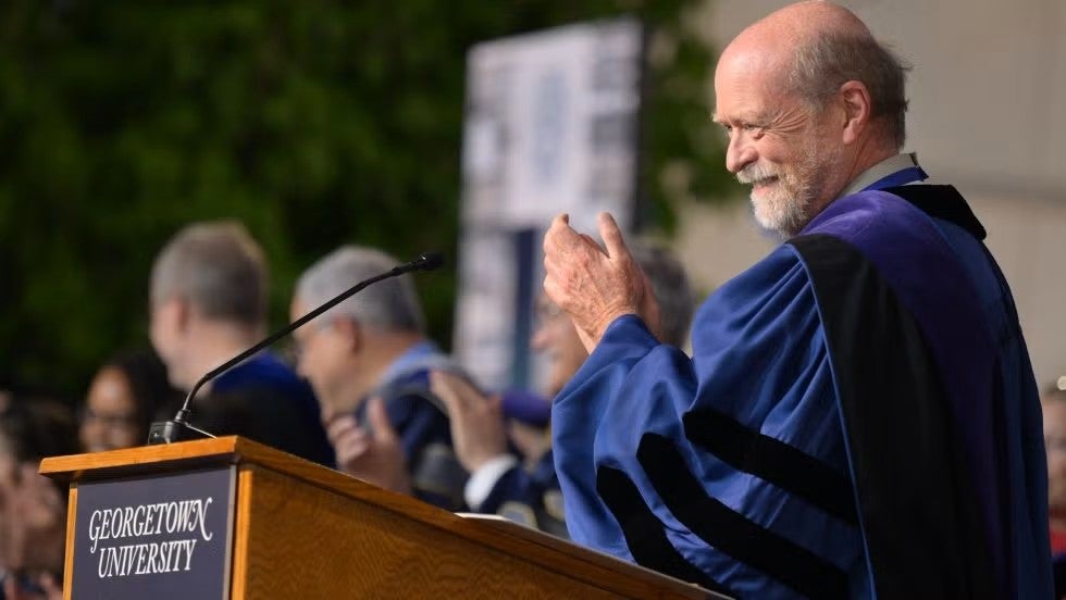 Dean William Treanor applauds behind a Georgetown University podium wearing academic regalia.