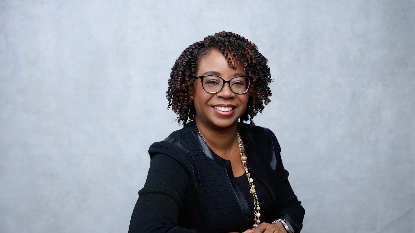 A woman smiles in front of a gray background. She wears glasses and a black blazer