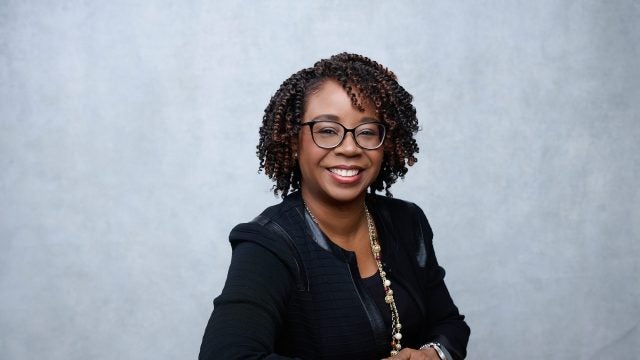 A woman smiles in front of a gray background. She wears glasses and a black blazer