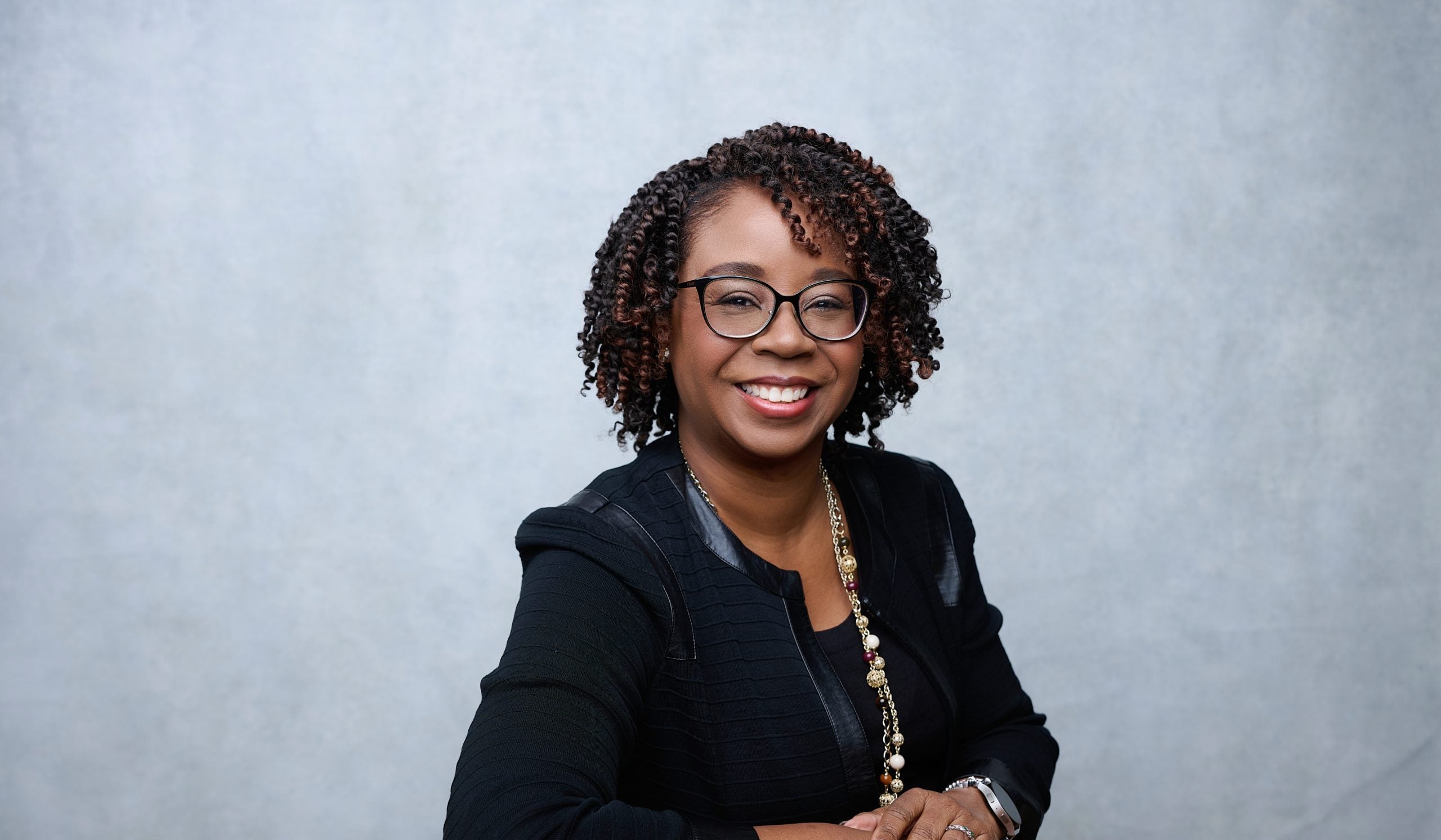A woman smiles in front of a gray background. She wears glasses and a black blazer