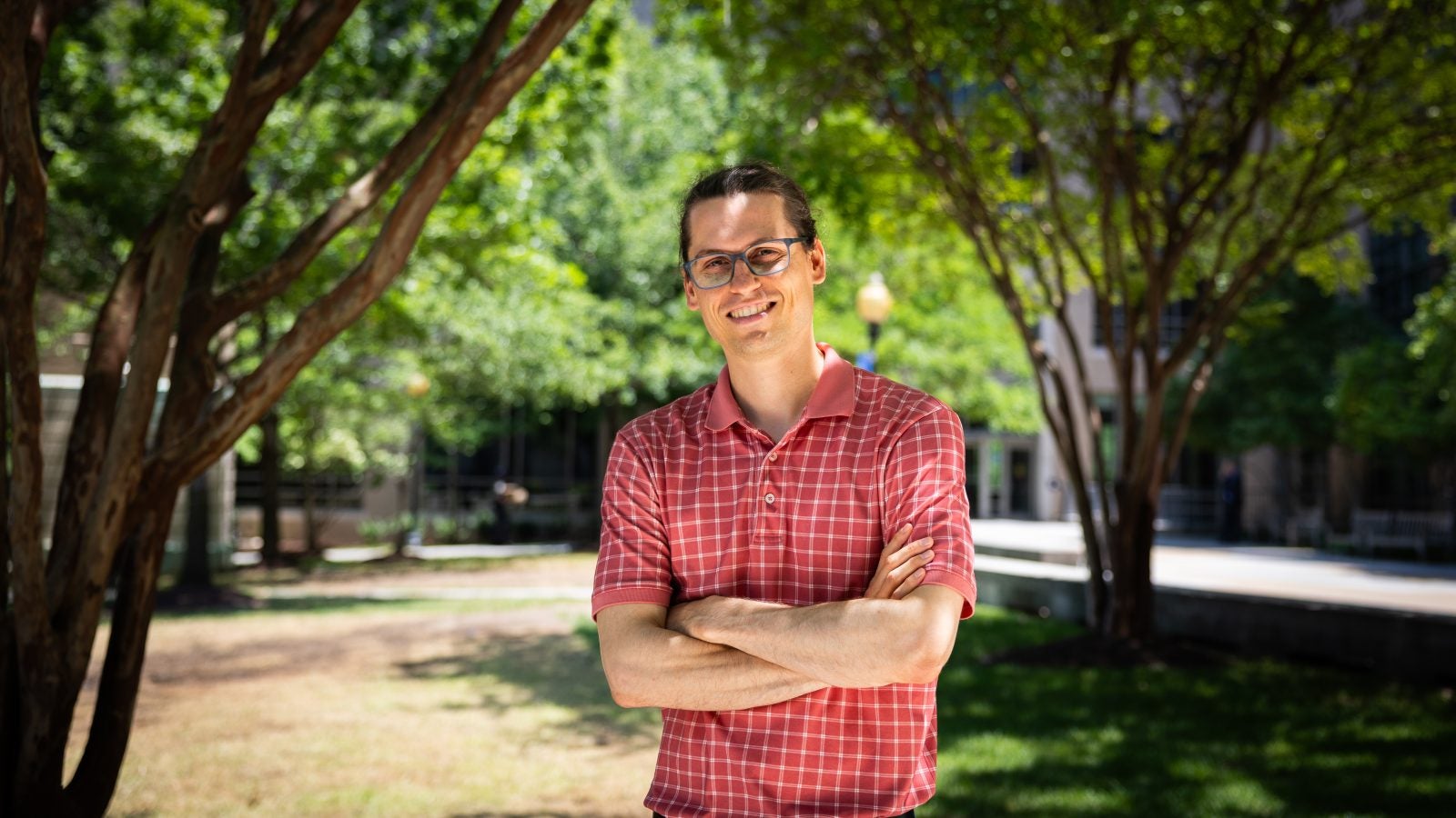 A man stands with his arms crossed in front of trees on a sunny day