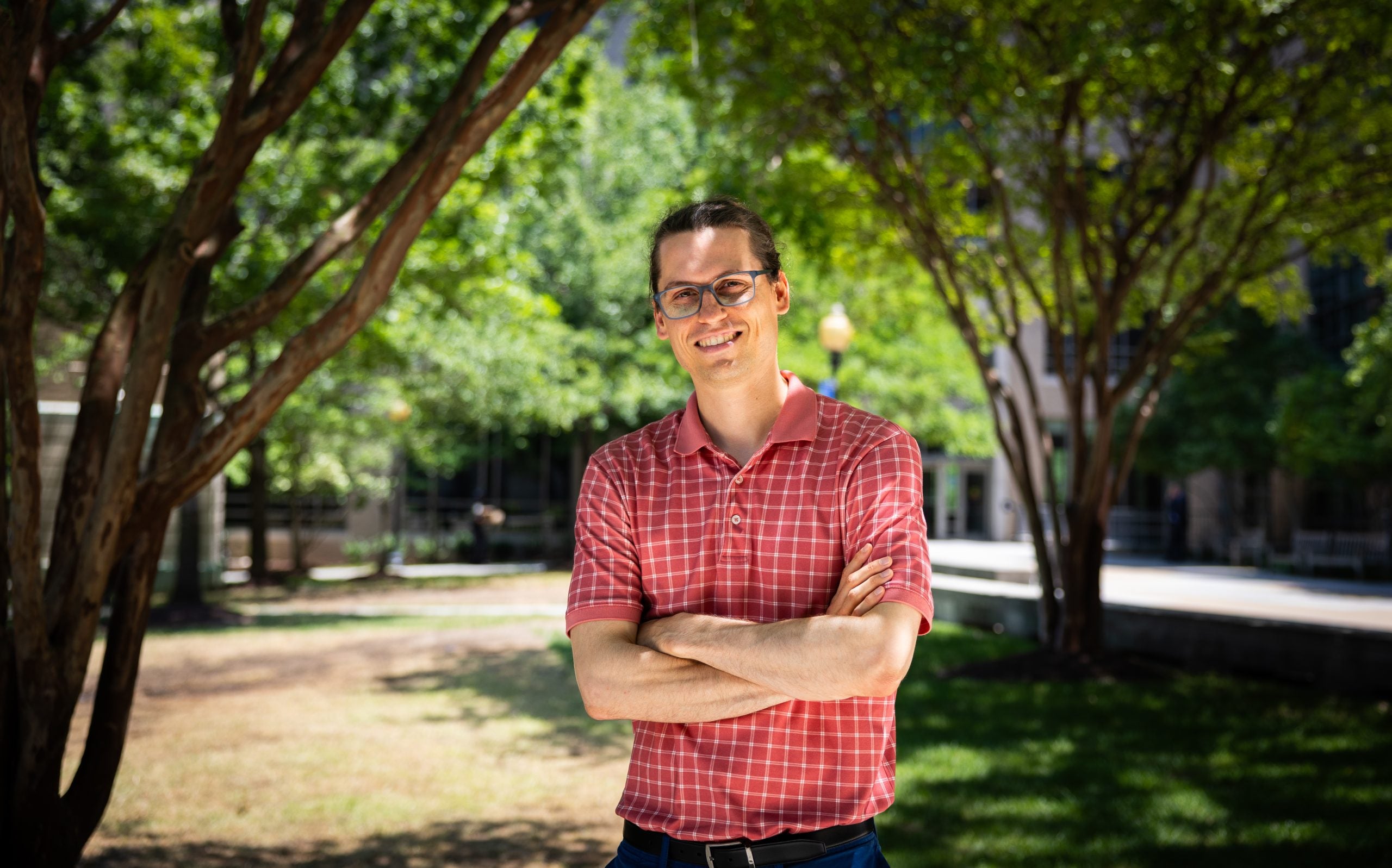 A man stands with his arms crossed in front of trees on a sunny day