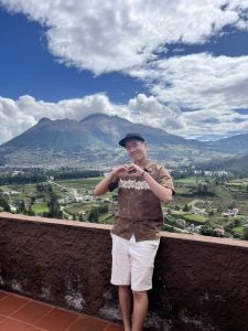 Jeremy wearing a cap holding his hands in a heart shape with a landscape of Ecuador behind him