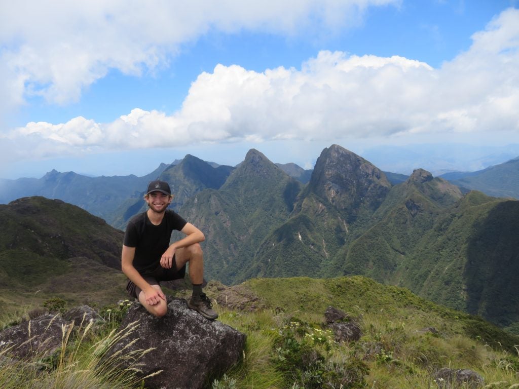 A male student perches on top of a mountain on a sunny day