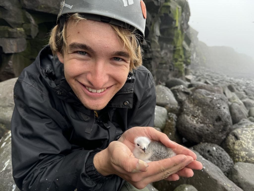 A male student holds a chick in his hands and smiles in front of a rocky shore.