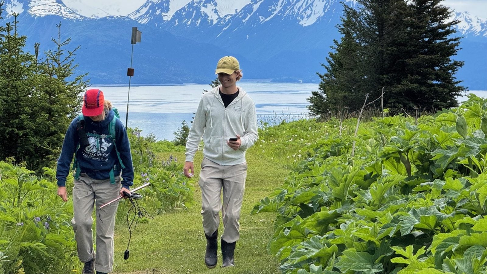 Two people walk on a grassy meadow in front of a bay and mountain behind them