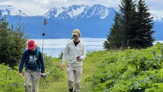 Two people walk on a grassy meadow in front of a bay and mountain behind them
