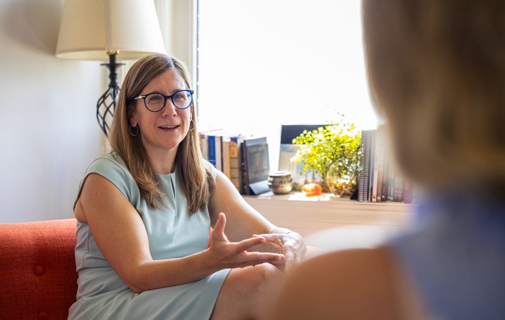 A woman with glasses and a light blue dress speaks with someone in front of a window while seated