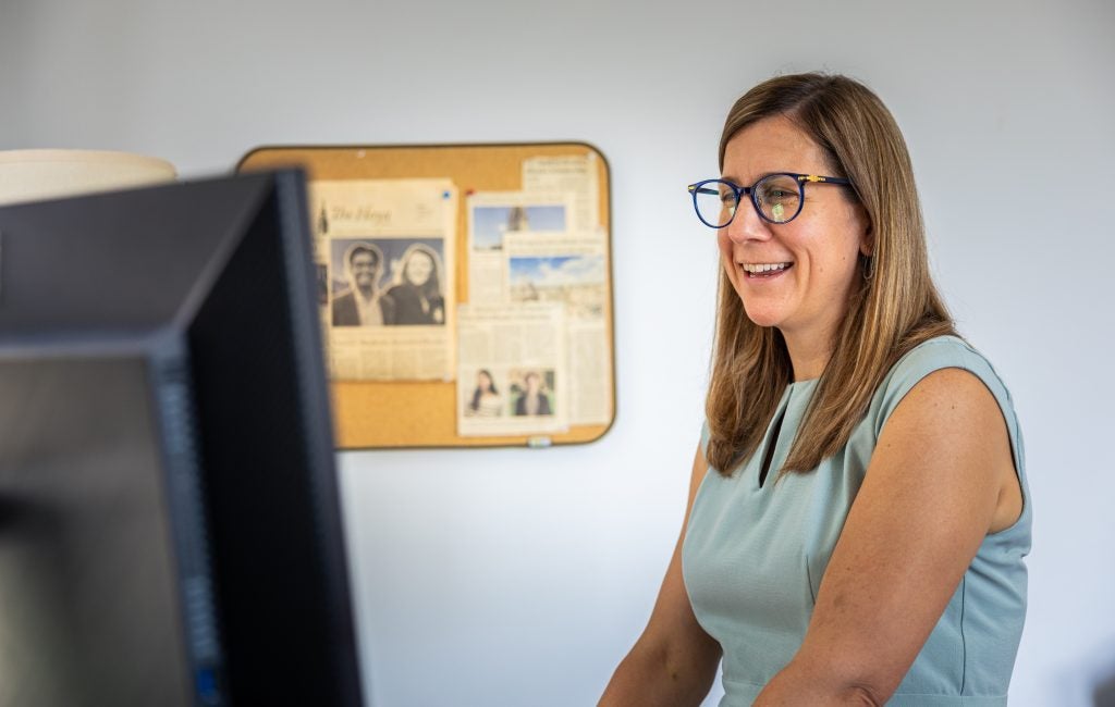 A woman wearing glasses and a light blue dress laughs while looking at her computer