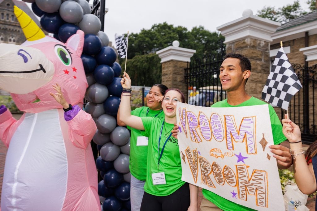 A student holds a sign that says "Vroom Vroom!" next to another cheering student and a student in a unicorn costume