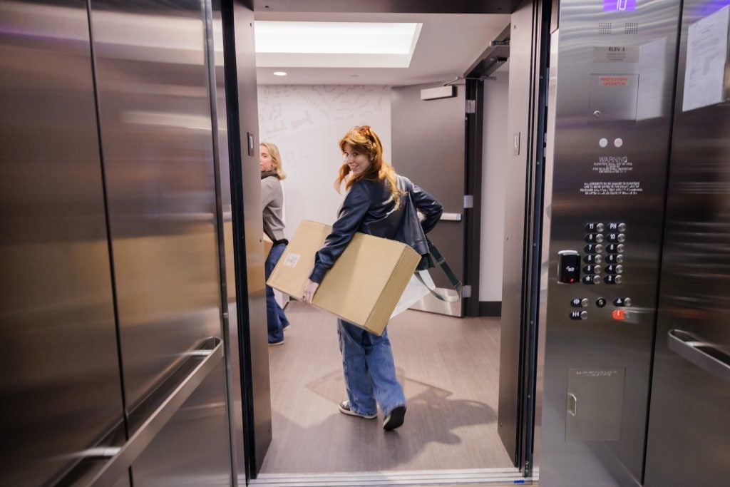 A student smiles while holding a rectangular box and getting off an elevator