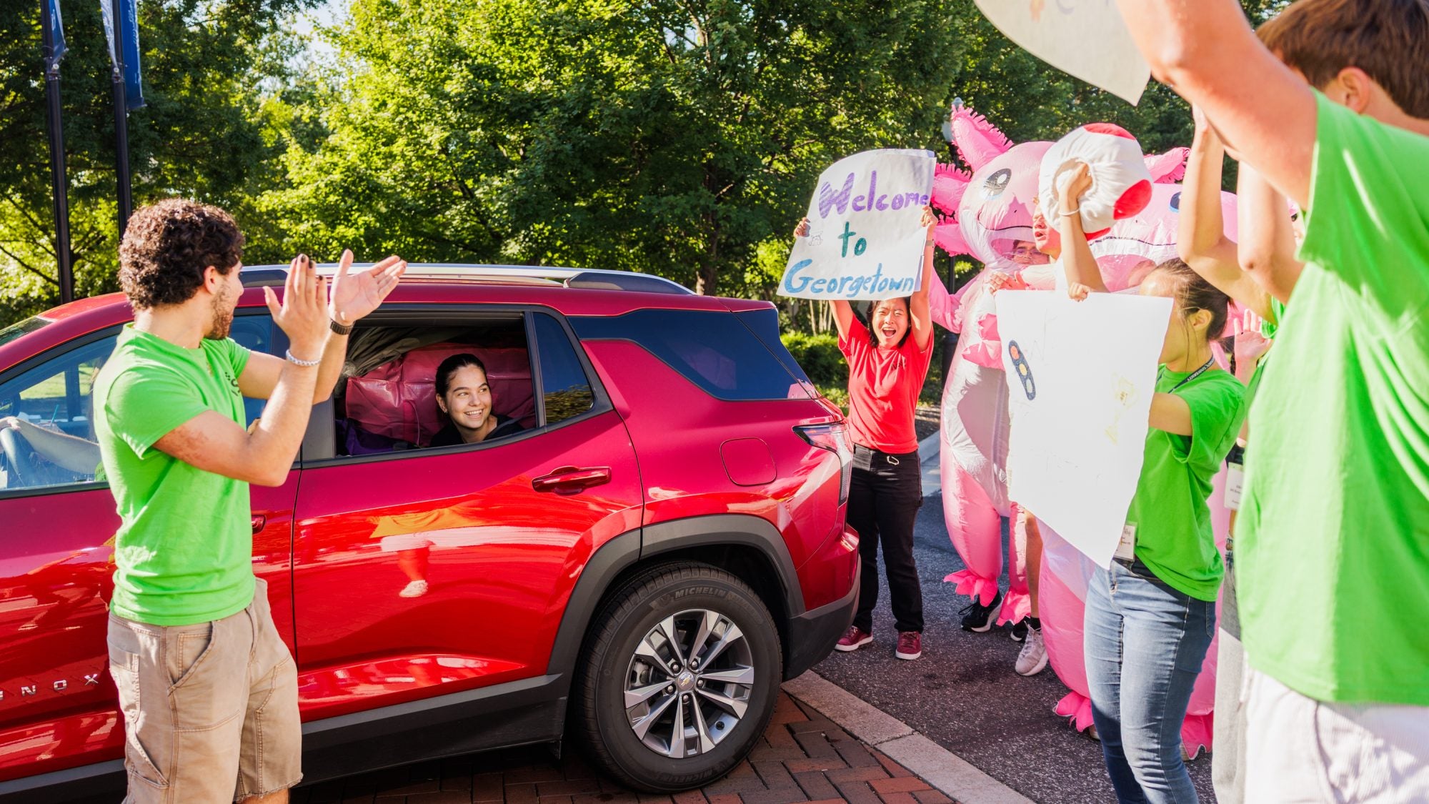 a student in a red car is welcomed by GU students in green shirts carrying welcome signs