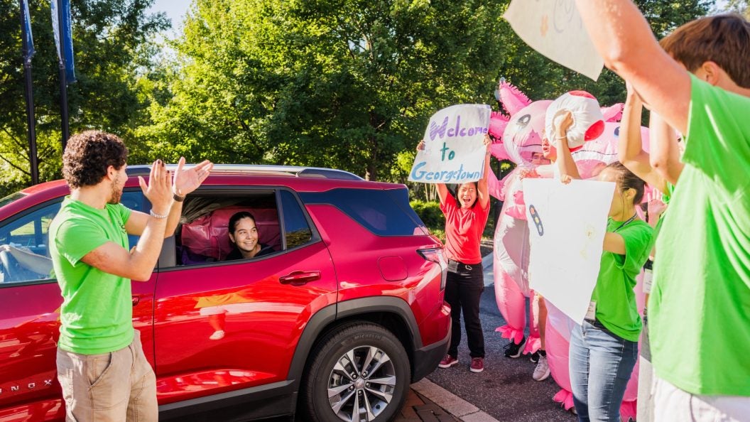 a student in a red car is welcomed by GU students in green shirts carrying welcome signs