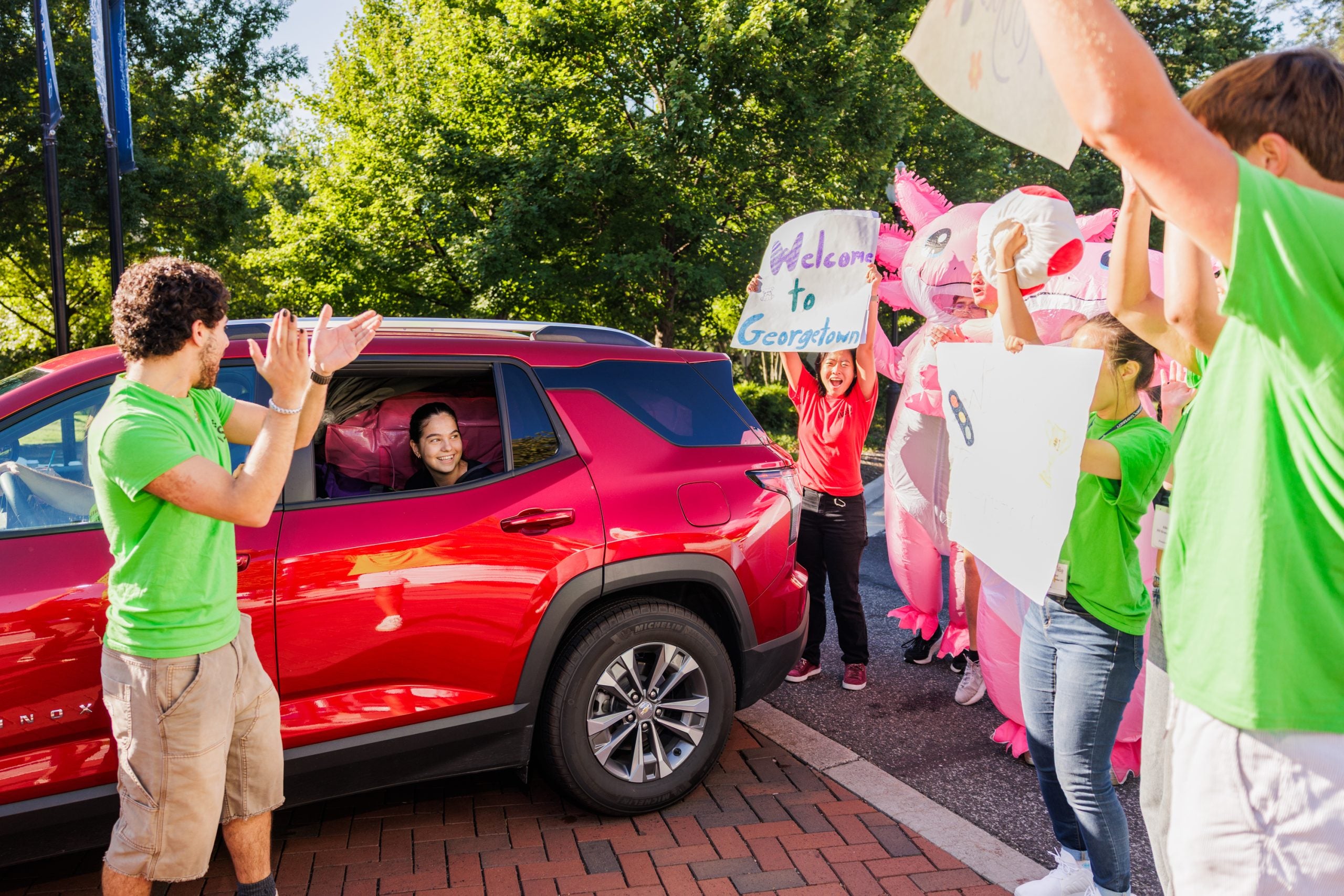 a student in a red car is welcomed by GU students in green shirts carrying welcome signs