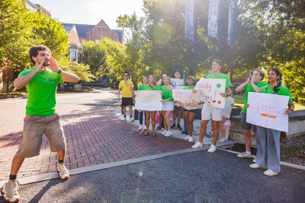 A male student raises his hands to his mouth in a cheer as other students hold signs and cheer