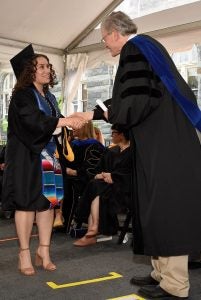 A student in a graduation gown and cap shakes a man's hand on her graduation day