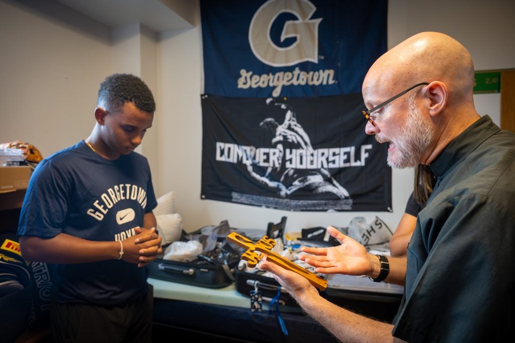 A Jesuit priest blesses a crucifix as student bows his head and prays