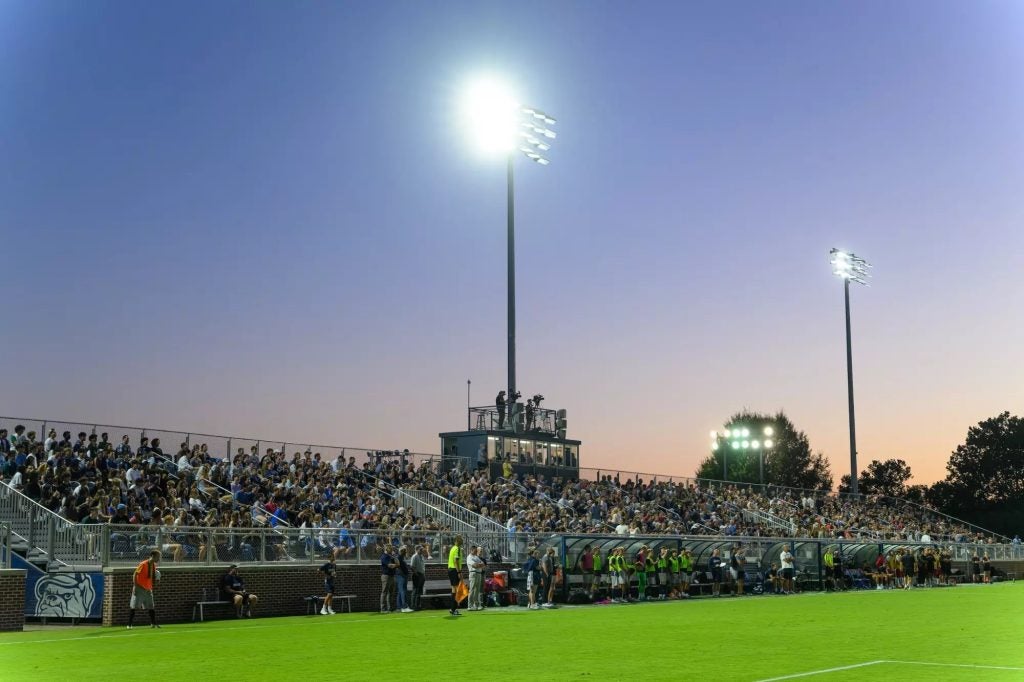 Stadium lights light up a soccer field as the sun sets