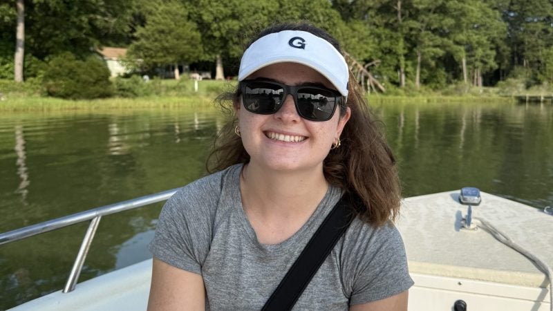 A student smiles on a boat with sunglasses and a cap.