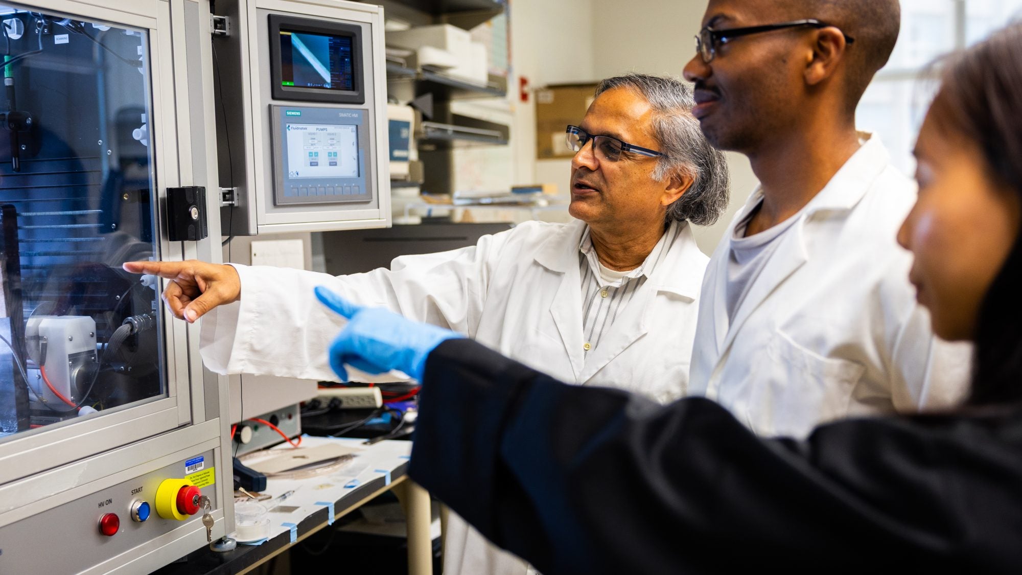 Mak Paranjape in a lab coat with two students looking at a lab device