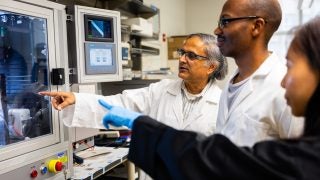 Mak Paranjape in a lab coat with two students looking at a lab device