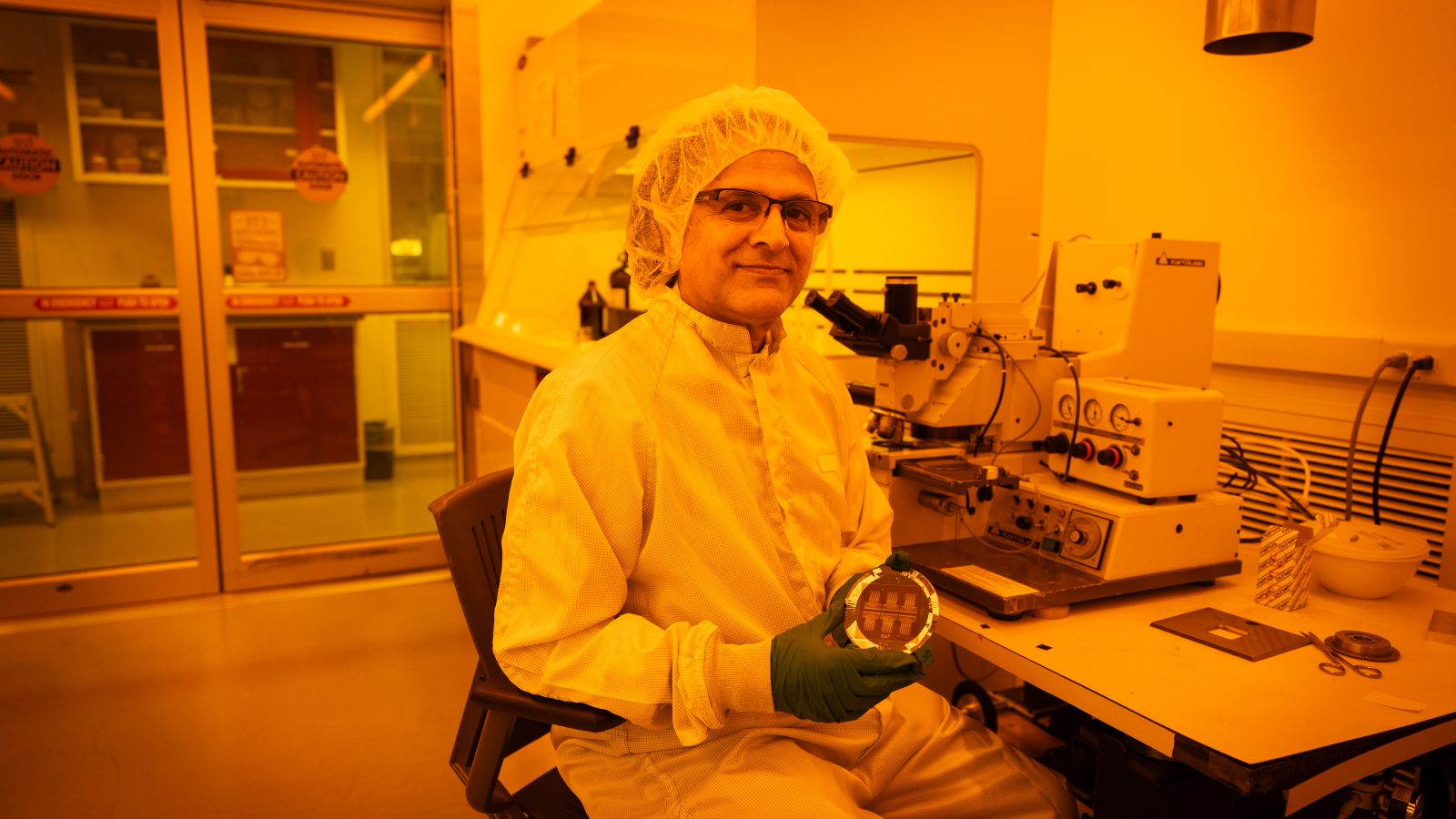 Mak Paranjape in a lab coat and a head cover holding a circular device in a yellow lit room