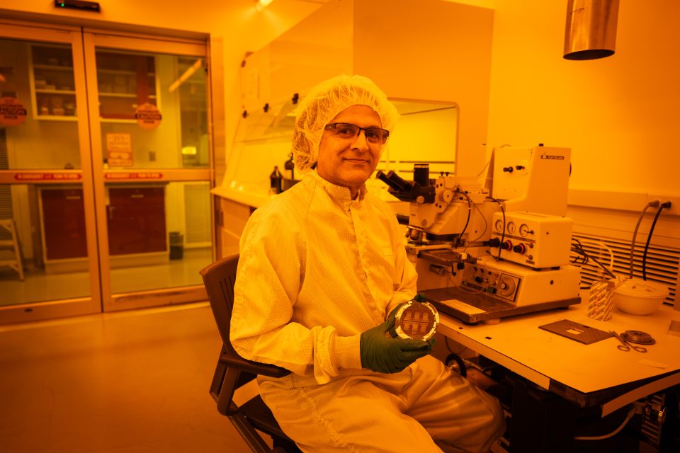 Mak Paranjape in a lab coat and a head cover holding a circular device in a yellow lit room