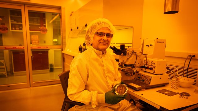 Mak Paranjape in a lab coat and a head cover holding a circular device in a yellow lit room
