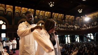 A professor helps a student put on a white coat on Gaston Hall&#039;s stage