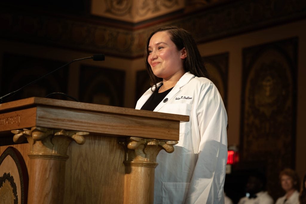 A woman smiles in a white lab coat with her name on the front while standing in front of a podium