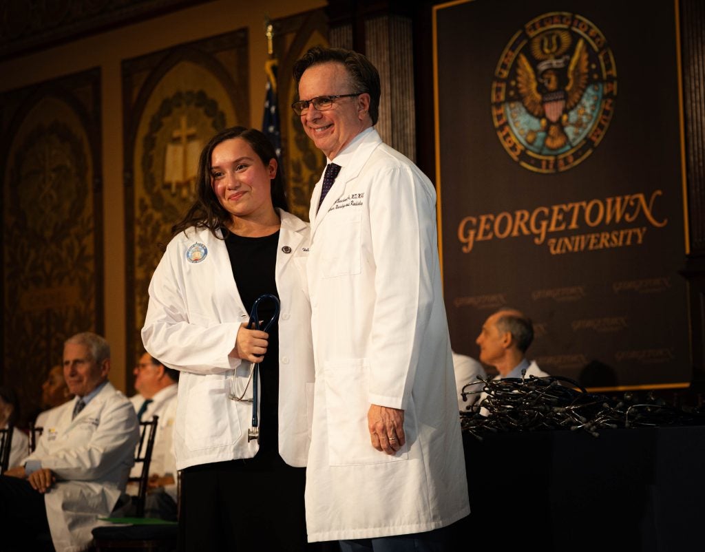 A student in a white coat poses with Dr. Norman Beauchamp, executive dean of the Georgetown School of Medicine, on Gaston Hall's stage 