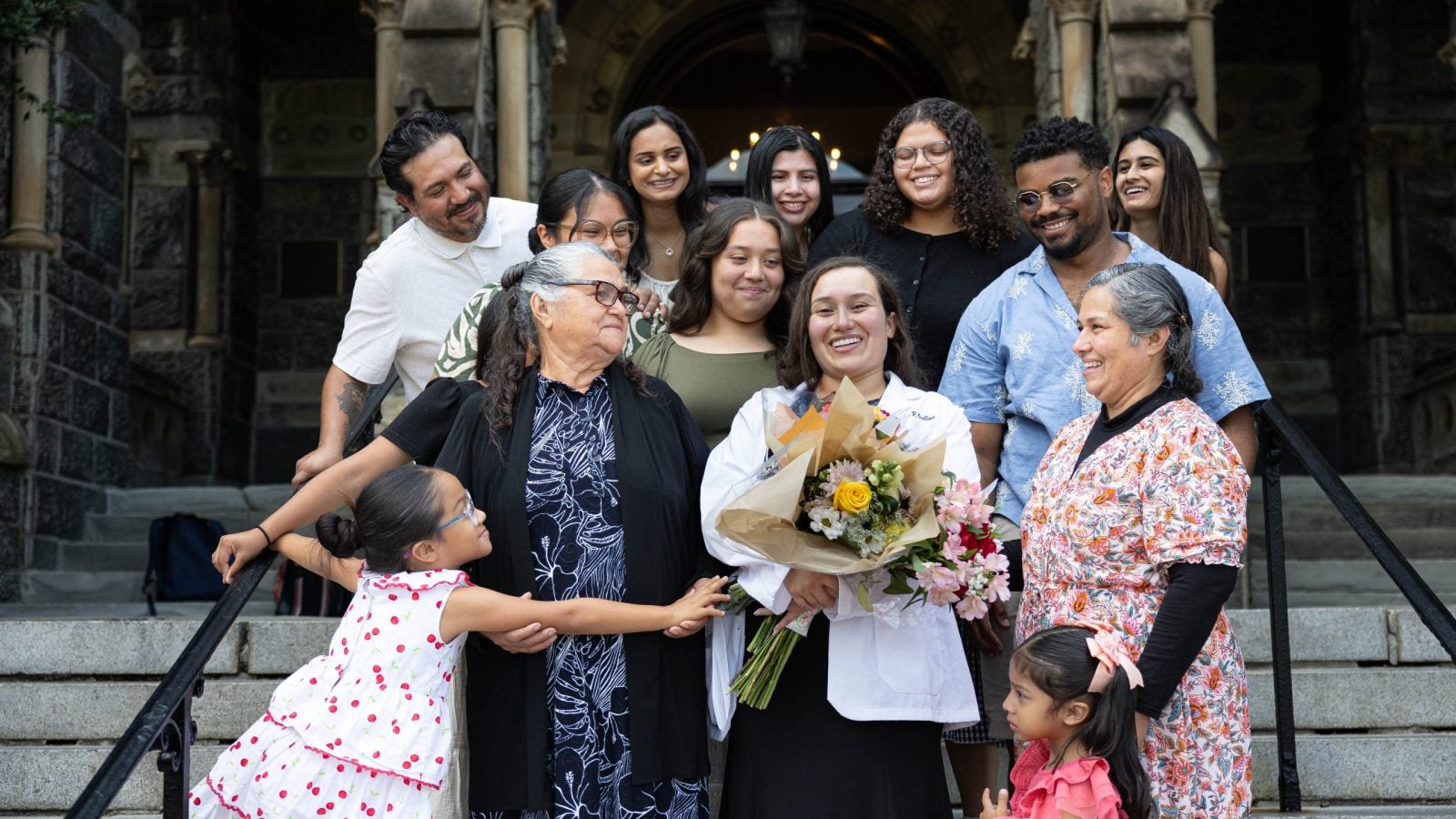 A family gathers around a student wearing a white coat and holding flowers on stone steps