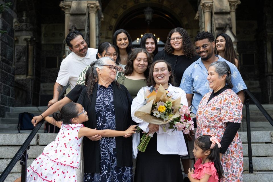 A family gathers around a student wearing a white coat and holding flowers on stone steps