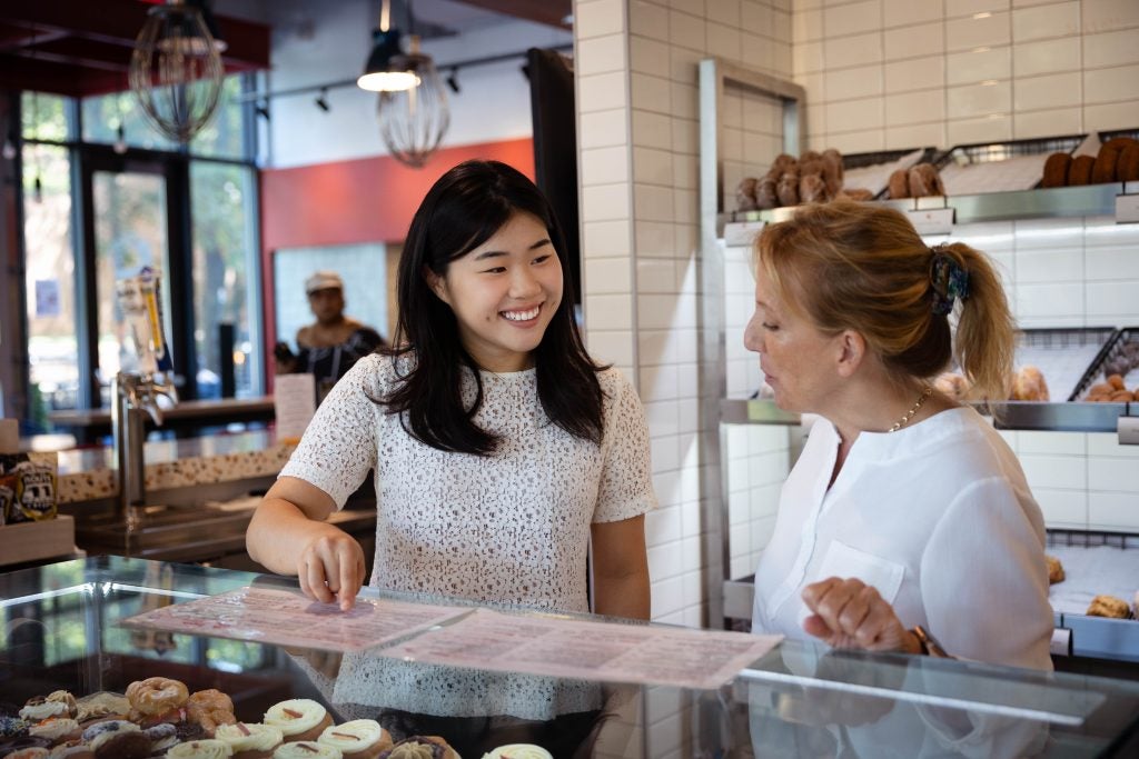 A student points and smiles at a menu at a donut shop to a woman