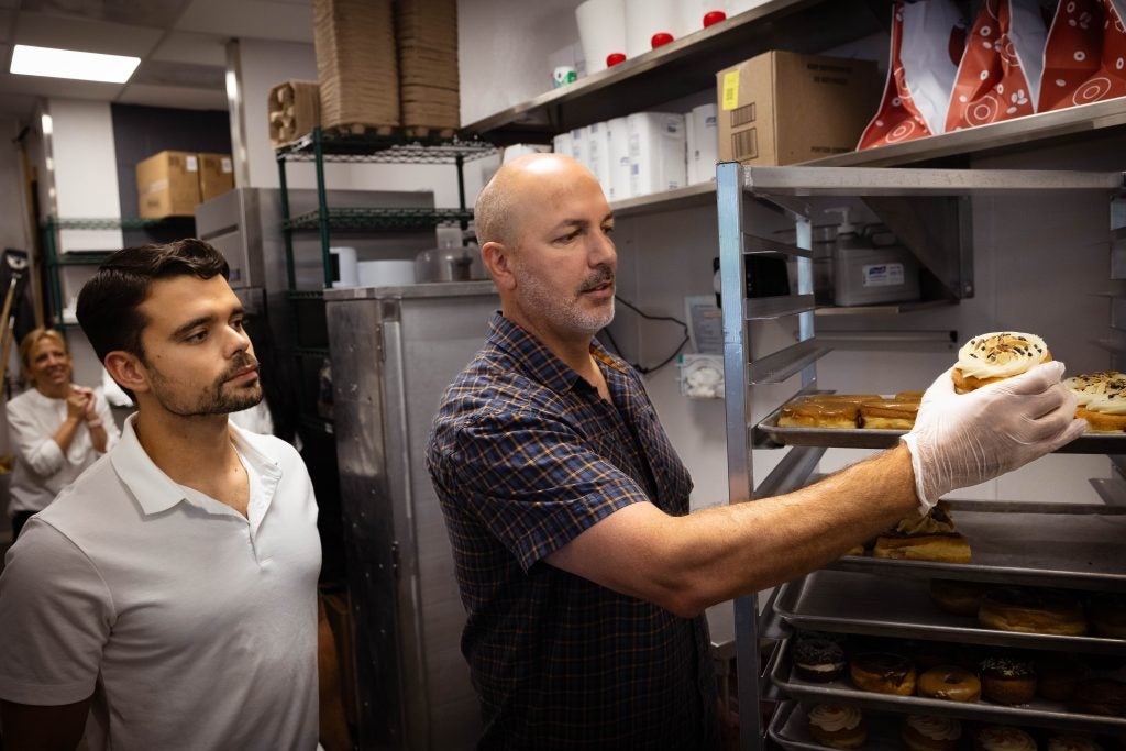 A man wearing plastic gloves picks up a donut off a baking tray while a student looks on