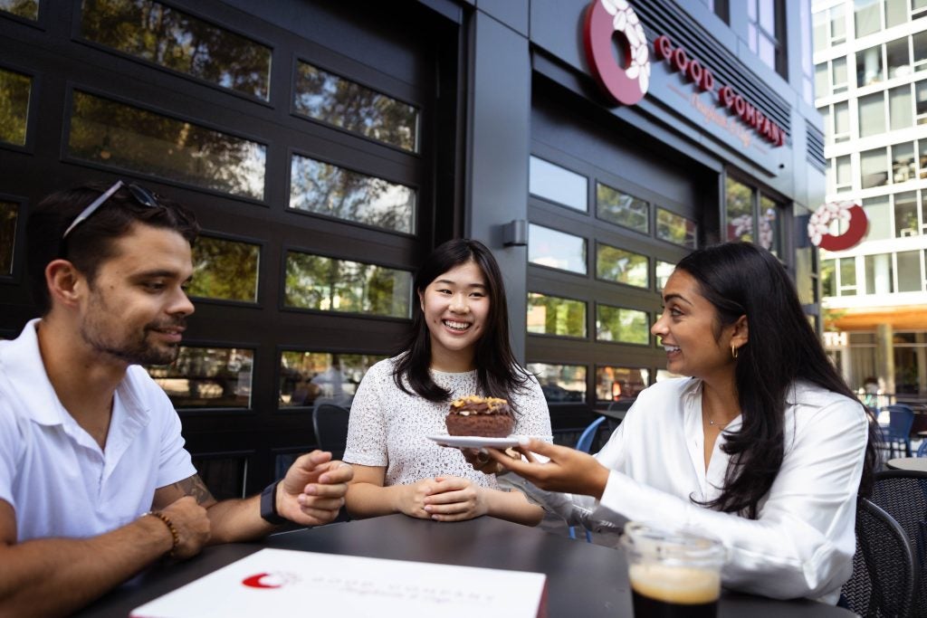 Two female students and a male enjoy a donut sitting on a patio outside a bakery