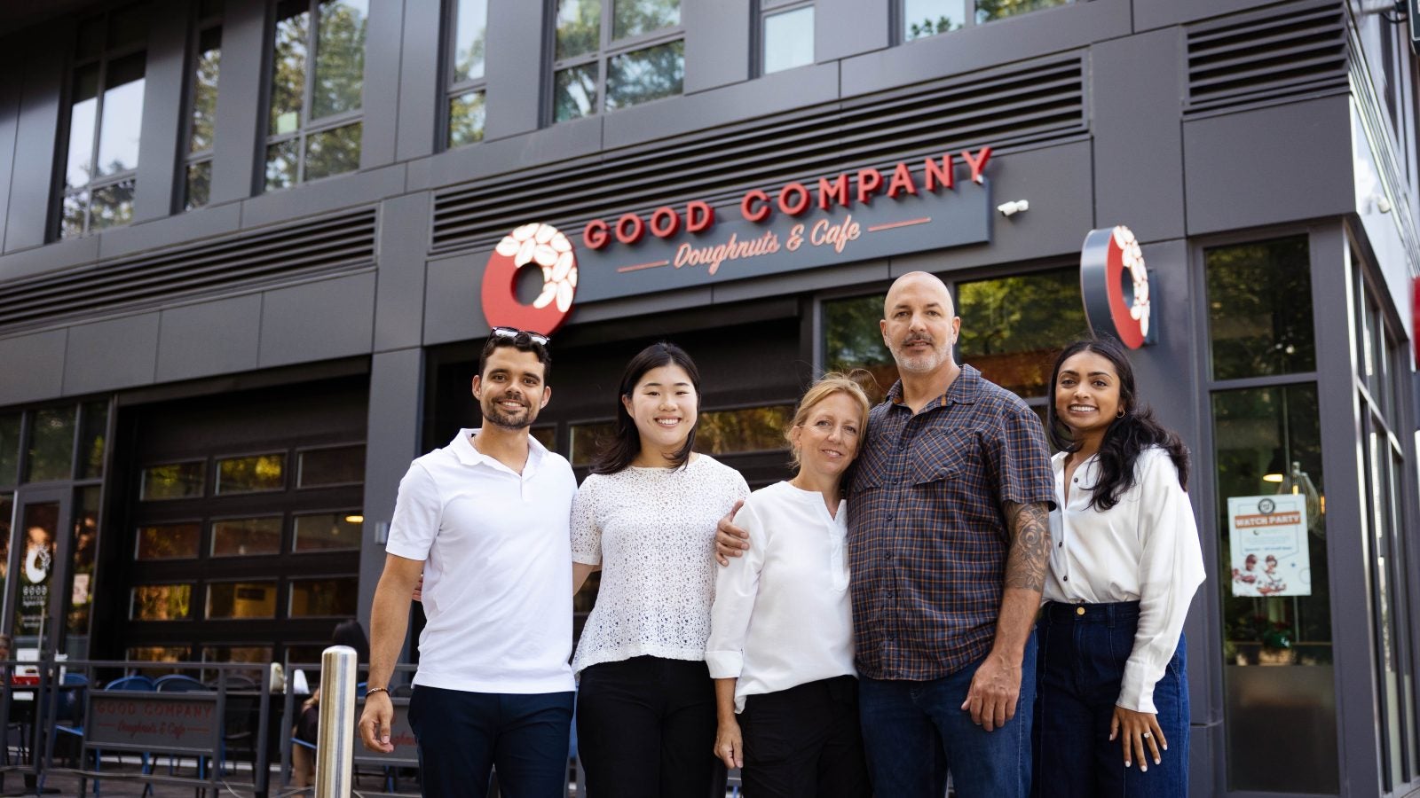 A group of students and owners of a bakery stand in front of a Good Company Doughnut &amp; Coffee cafe