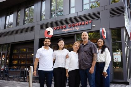 A group of students and owners of a bakery stand in front of a Good Company Doughnut &amp; Coffee cafe