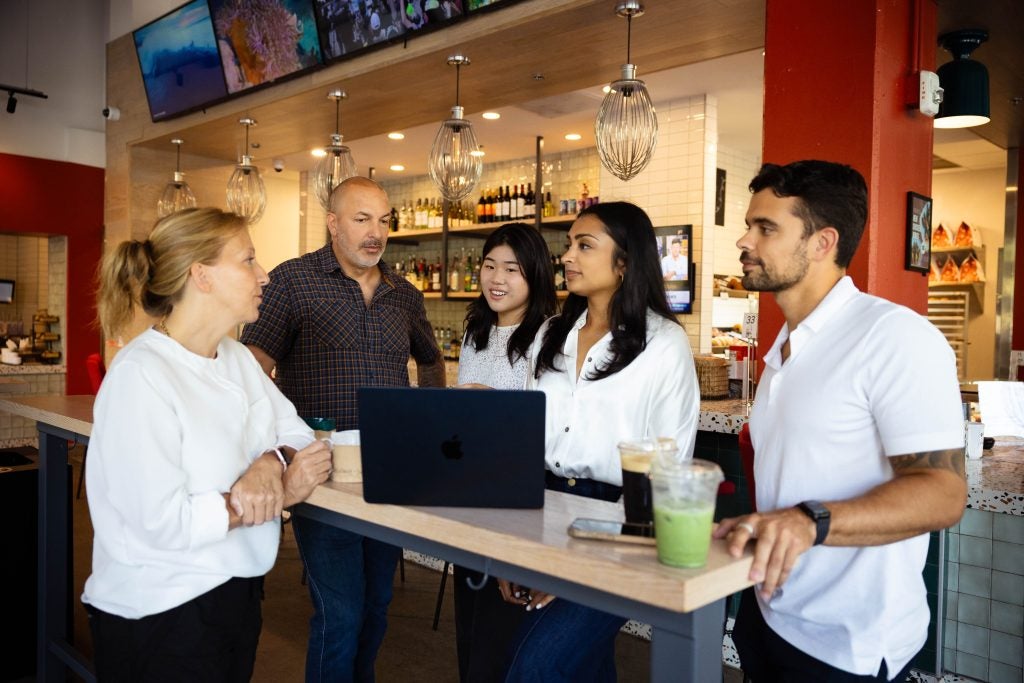 A group of students consult with the owners of a bakery shop while standing around a table