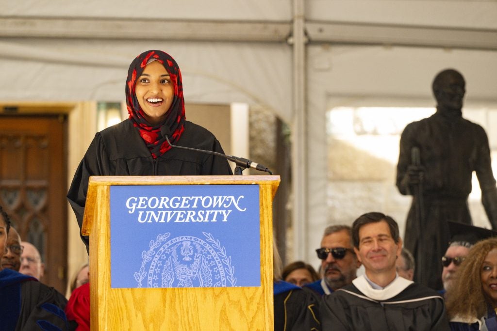 A woman wearing a hijab and a black graduation gown smiles from behind a podium