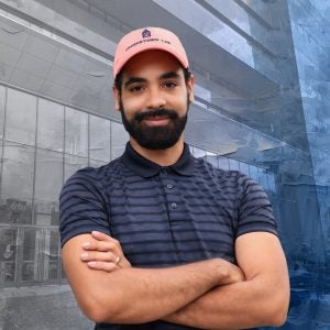 A man in a orange baseball cap stands in front of a textured background of a glass building
