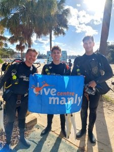 Three young men in diving gear holding a sign on a sunny day