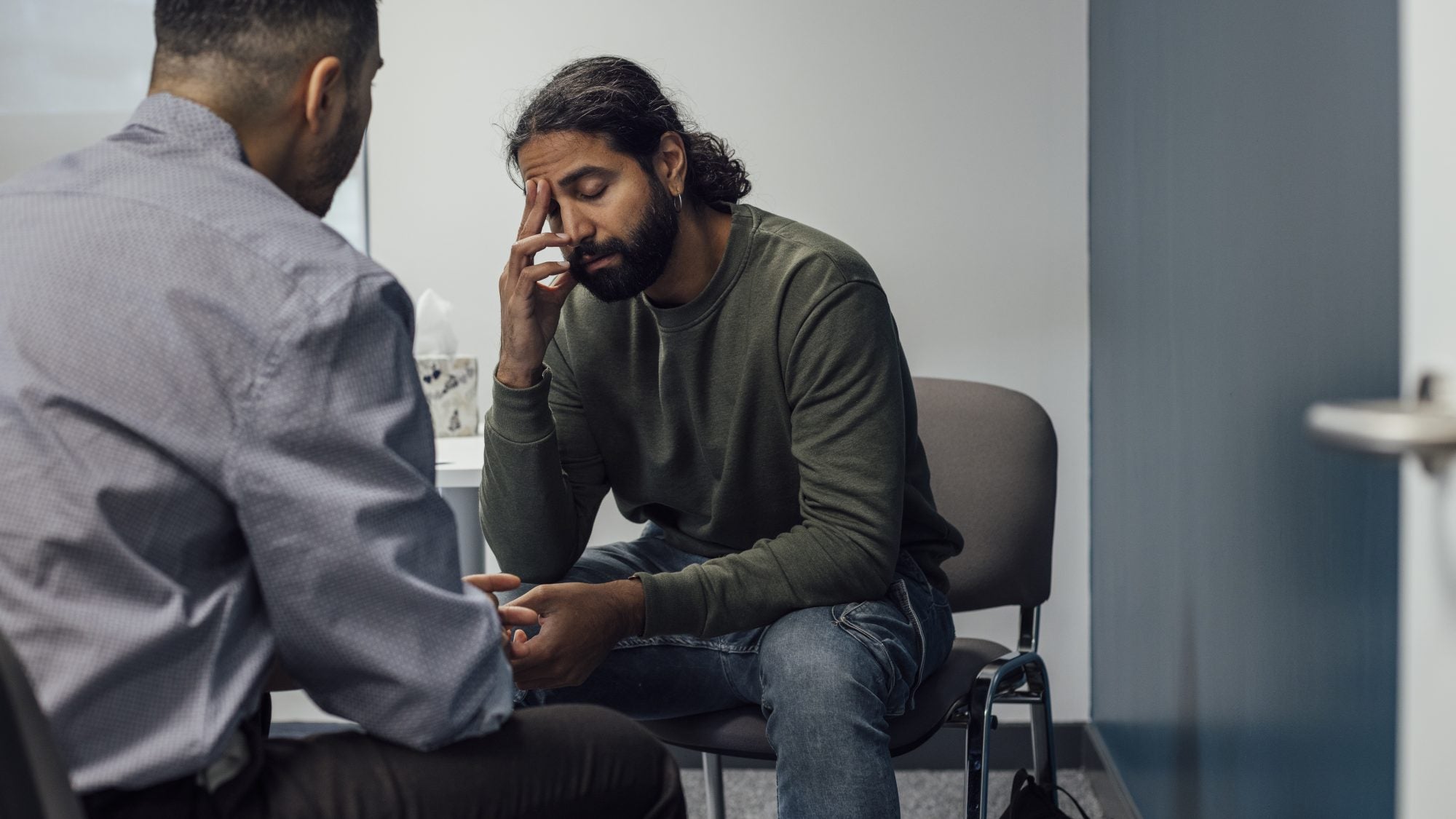 Over-the-shoulder shot of a doctor and man. The man looks sad and depressed after hearing news from the doctor. The man&#039;s eyes are closed and has one hand covering part of his face.